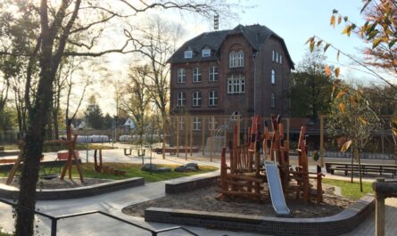 Ein neugebauter Spielplatz und Treffpunkt für das Viertel Georgswerder. Im Vordergrund ist ein Spielgerät mit Rutsche, im Hintergrund das historische Schulgebäude aus Backstein. Links im Bild sind Bäume und Sonnenlicht zu sehen.