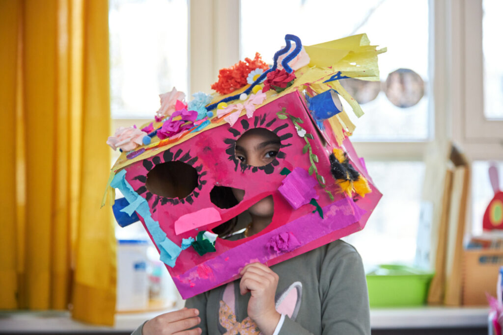Eine Vorschülerin, die ihre rosane, bunt beklebte Pappkarton-Maske aufgesetzt hat und durch die ausgeschnittenen Augen guckt.