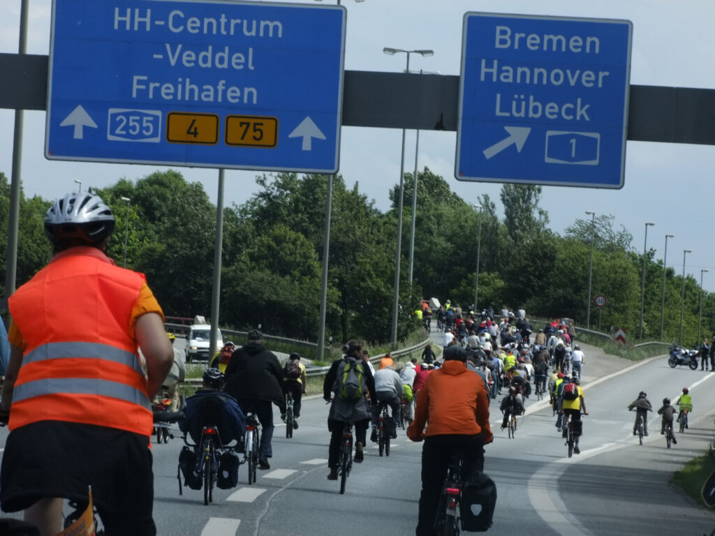 Ein großer Pulk Fahrradfahrer:innen, Alt und Jung, mit bunten Helmen und Jacken, fährt über eine Autobahn. Die Radler:innen sind von hinten aufgenommen.