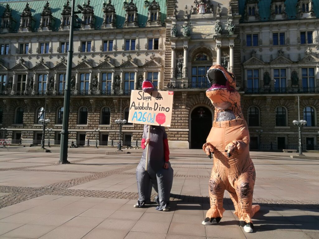 Auf dem Platz vor dem Hamburger Rathaus stehen zwei als Dinosaurier verkleidete Personen. Einer hält ein Schild hoch, auf dem steht: "Stoppt den Autobahn-Dino A26 Ost!"