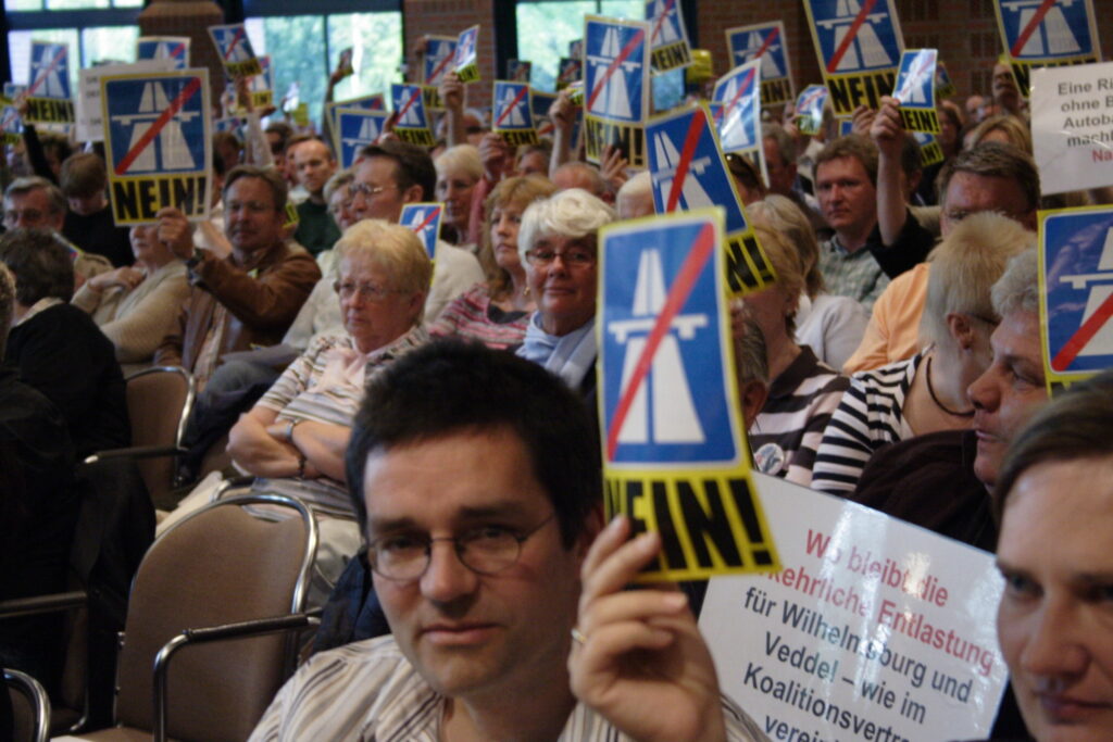 Die Stuhlreihen eines Veranstaltungssaals sind dicht besetzt mit Menschen, ein großes Publikum. Viele der Menschen halten ein Schild hoch, darauf ist eine durchgestrichene Autobahn abgebildet, unten steht in Großbuchstaben "NEIN!". Es ist ein Schilderwald in diesem Publikum.