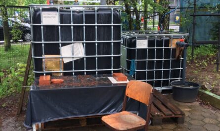 Frontalansicht Biogasanlage, zwei schwarze Container mit Gittergehäuse. Aufgestellt draußen auf einem Tisch, vor der Anlage ein Stuhl. Einfülltrichter für Bioabfälle auf dem Container.