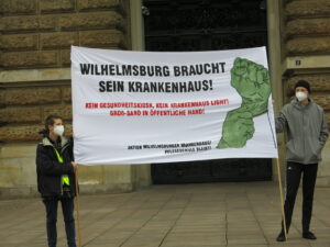 Zwei Demonstranten stehen vor dem Rathaus mit einem Transparent: Wilhelmsburg braucht sein Krankenhaus 