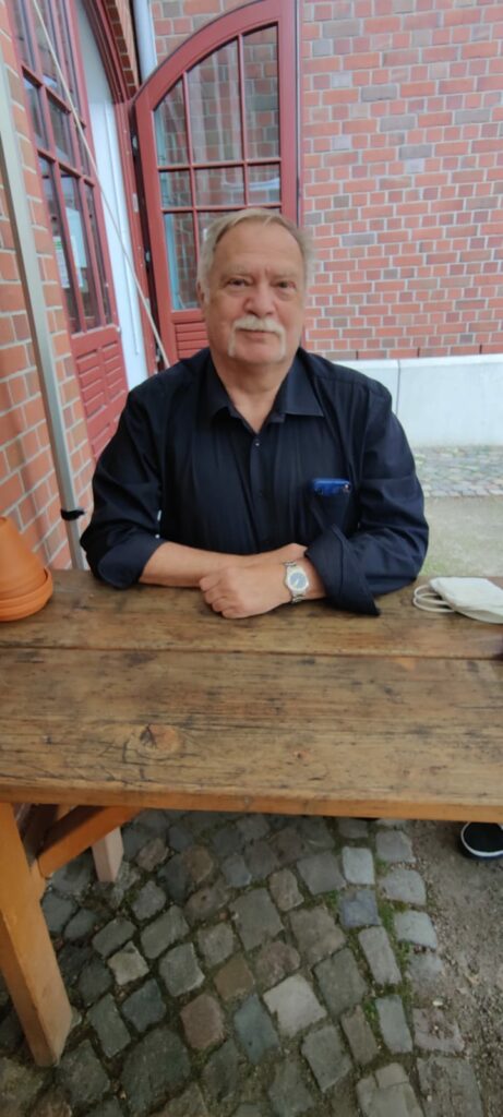 Dieter Thal, ein verhalten lächelnder Mann mit Schnauzbart, an einem Holztisch im Innenhof der Ballinstadt.