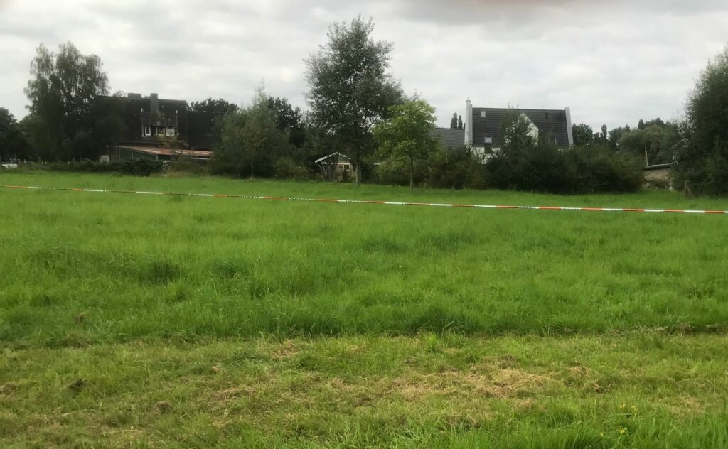 Im Verrdergrund eine grüne Wiese. Dahinter Bäume, Büsche und Häuser. In der Mitte ein Flatterband.
