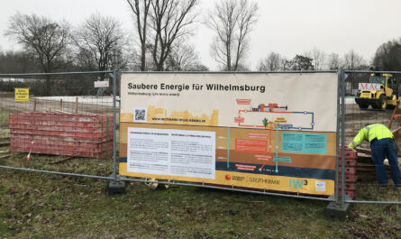 Um das Gelände der Geothermieanlage. Davon ist ein Teilstück zusehen mit dem Banner "Saubere Energie für Wilhelmsburg" und einer Beschreibung, die auf dem Bild aber zu klein ist, um sie lesen zu können. rechts sieht man einen Bauarbeiter und ein kleines Baufahrzeug mit dem Namen MAC und einige Bäume sind auch auf dem Bild.