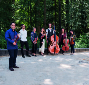 Sieben Musiker:innen des Hamburg Stage Ensembles stehen mit ihren Instrumenten auf einem Platz vor einem Wald
