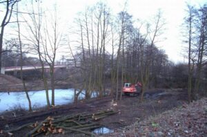 Hinten links in der Mitte eine Brücke, davor ein größerer Teich an dessen Ufer im Vordergrund Bäume und davor liegen einige abgeholzte Bäume. In der Mitte ein rotes Baufahrzeug.