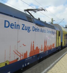 Eine Metronom-Lokomotive am Bahnsteig mit der Aufschrift: Dein Zug. Dein Auto. deine Region
