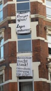 Ein mehrstöckiges Eck-Backsteinhaus. Daran zwei Banner: Auf einem steht: "Grenzen öffnen, Lager evakuieren", auf dem anderen Rassismus macht krank, Gesundheitsversorgung für Alle!"