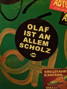 Auf grünem, bemaltem Hintergrund klebt ein runder, schwarzer Sticker. Darauf ist in gelben Großbuchstaben geschrieben: "Olaf ist an allem Scholz."