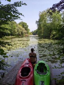 im Vordergrund liegen zwei Kajaks auf einem Steg, eineweiblich gelesene Person sitz mit dem Rücken zum Betrachtenden davor. Blick aufs Wasser, ein Fluss, von Bäumen umringt.