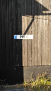 An einer Holzwand ist ein KfZ-Schild angebracht. Links darauf ein weißes P auf blauem Grund, auf dem Rest des Schildes in schwarzen Großbuchstaben auf weißem Grund das Wort "Presi".