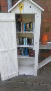 Ein weiß gestrichenes Holzhäuschen, die Tür steht offen, darin stehen Bücher.