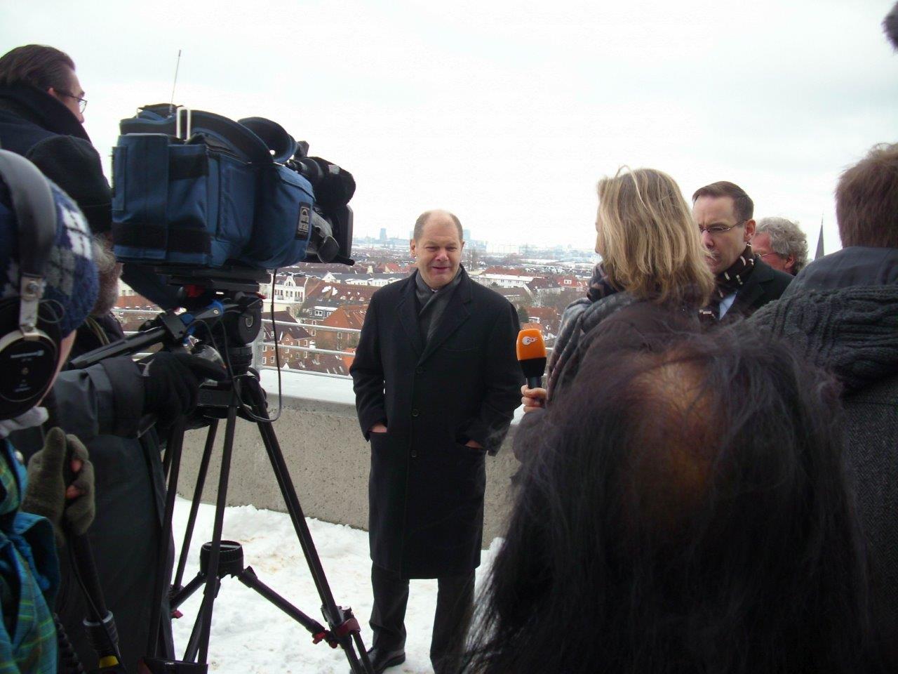 Im Vordergrund einige Journalist:innen, die mit dem Rücken zur Kamera Olaf Scholz interviewen und filmen. Im Hintergrund der Ausblick vom Bunker in der Neuhöfer Straße
