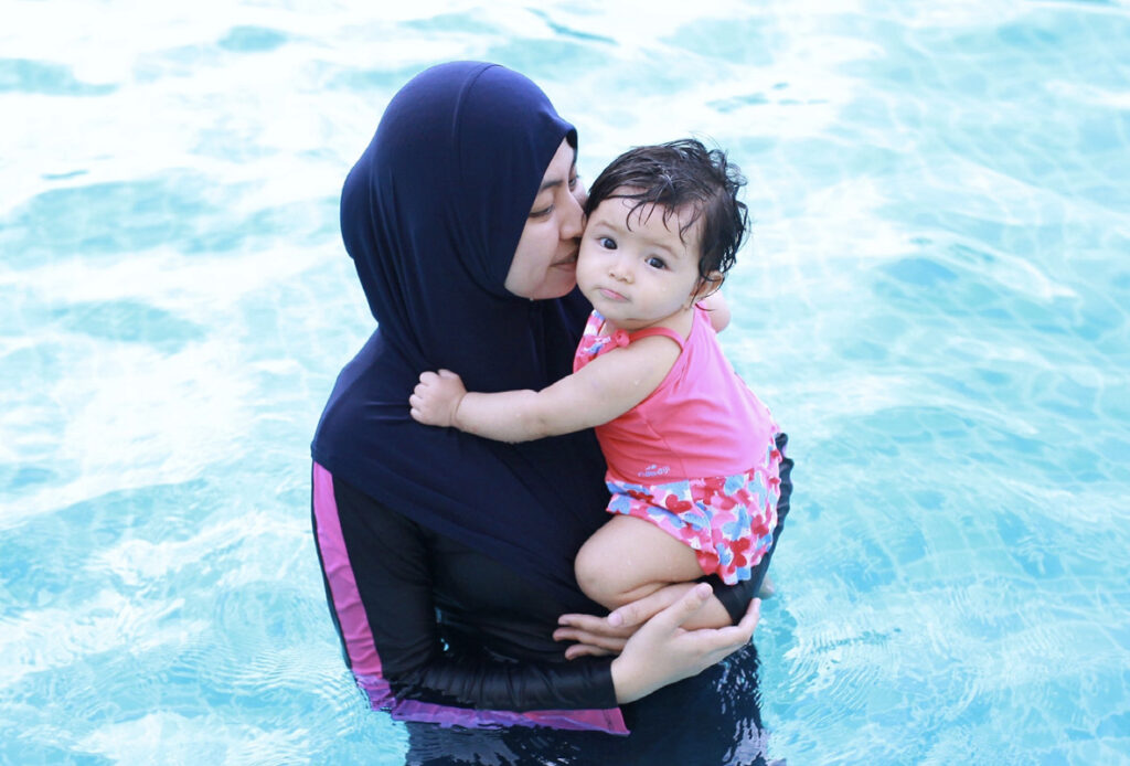 Frau im Burkini steht im Wasser und hält ein Baby auf dem Arm