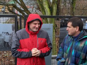 Zwei Männer, einer in roter Jacke mit Kapuze, der andere mit kurzen dunklen Haaren und einem bunten Mantel stehen vor einem Zaun, sehen sich an und Lächeln