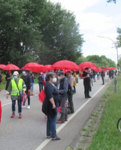 Auf der Kornweide stehen viele Menschen in langer Reihe mit aufgespannten roten Regenschirmen