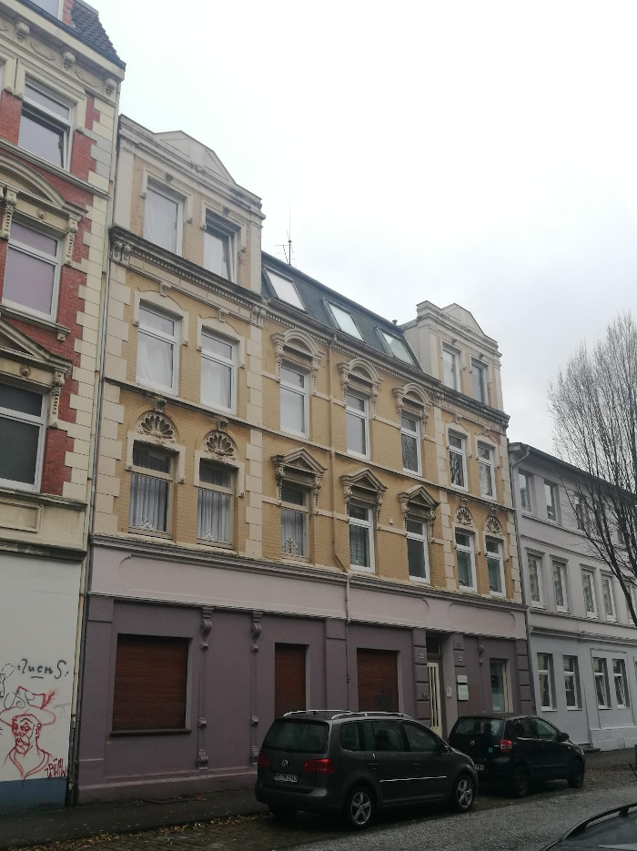 Ein vierstöckiges Haus. Anfang des 20. Jahrhunderts gebaut mit viel Stuck über den Fenstern. Die mittleren zwei Etagen gelb. Das Erdgeschoss, der Gewerbebereich ist mauve. 