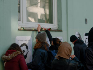 Menschen legen Blumen auf einen Fenstersims. Darunter eine Wand, auf der Plakate mit den Namen und Gesichtern der Ermordeten des Anschlags zu sehen sind.
