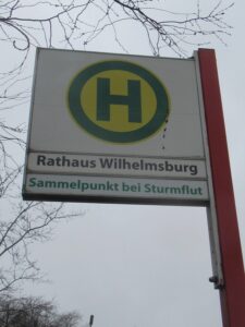 Das Haltestellenschild Rathaus Wilhelmsburg mit dem grünen Zusatz: Sammelpunkt bei Sturmflut