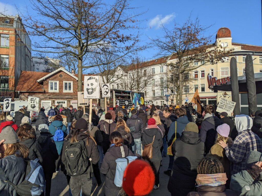 Eine Menschenmenge steht mit Blickrichtung zu einem Lautsprecherwagen auf dem Stübenplatz. Einige tragen Schilder mit den Gesichtern der Ermordeten.