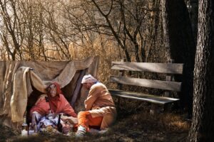 In einem Wald ist ein notdürftiges Zelt vor einer Bank aufgebaut. Davor ein Paar, das offensichtlich obdachlos ist in ärmlicher Kleidung. 