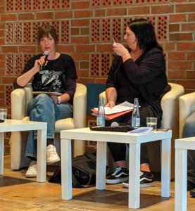 Sophie Brüll (links) und Nathalie Meyer (rechts), sitzen auf cremfarbenen Sesseln auf dem Podium. Brüll sprichts ins Mikro, Meyer sieht zu ihr und hebt einen Stift.