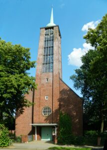 Vor einem tiefblauen Himmel ist die Vorderseite der Kirche zu sehen mit dem markanten quadratischen Turm. In der oberen Hälfte sind Glasscheiben eingesetzt. Links und rechts im Bild dicht belaubte Bäume.