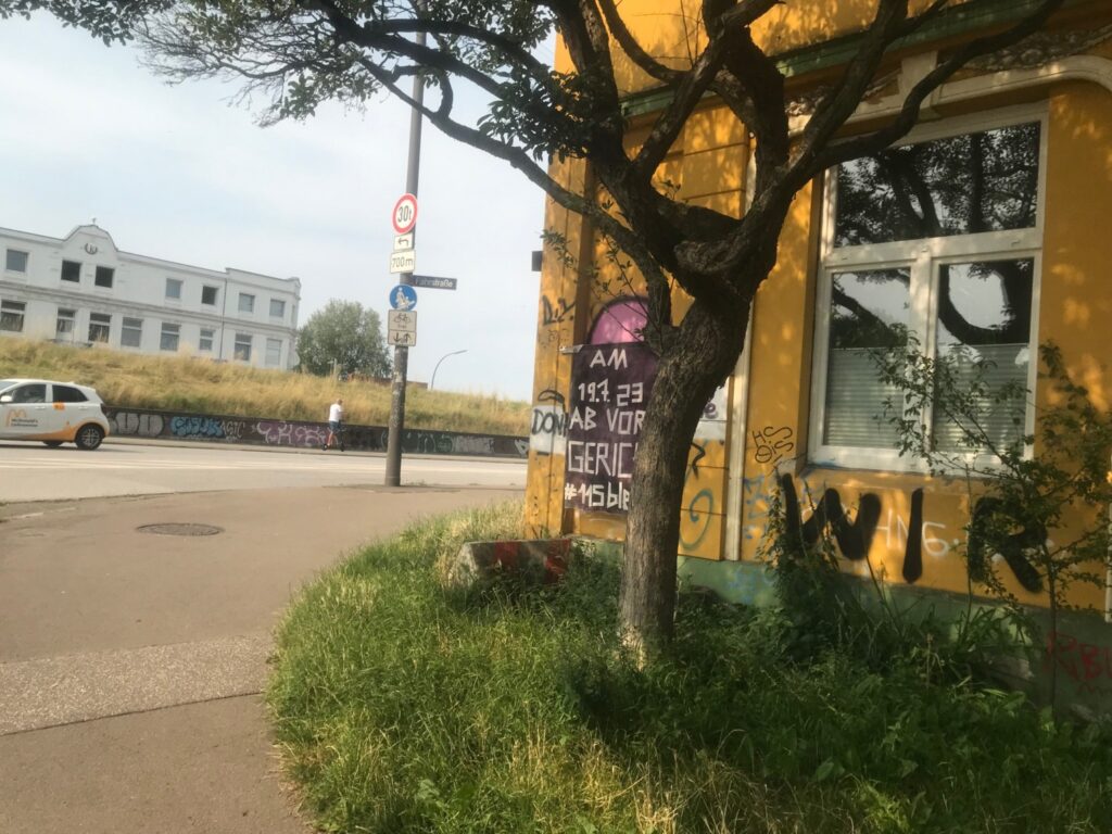 Eine Häuserecke mit einem gelben Haus, ein Baum davor, ggü. ein Deich. An dem Haus ein Schild, auf dem steht "am 19.7. ab vor Gericht, 115 bleibt!"