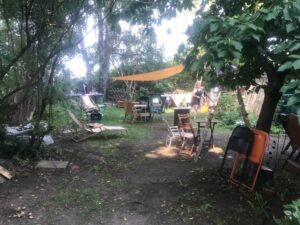 Vorbereitung für ein Infocafé im Garten. Überall stehen kleine Sitzgruppen und Liegestühle. In der Mitte ist ein gelbes Sonnensegel gespannt und ringsum stehen Bäume und Büsche.