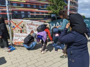 Mehrere Frauen befestigen Transparente an Straßenlaternen. Darauf ist zu lesen: "Wohnungen für Alle, raus aus den Camps!". Im Hintergrund die Fassader der Behörde für Stadtentwicklung und Wohnen.