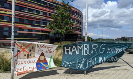 Im Hintergrund die Behörde für Stadtentwicklung und Wohnen, im Vordergrund 2 aufgehängte Transparente. Auf einem steht: Wohnungen für alle! Raus aus den Camps!" auf dem anderen "Hamburg, ich brauche eine Wohnung von dir!"