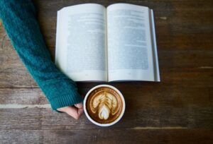 Symbolbild: Ein aufgeschlagenes Buch, ein Arm im Wollpulli greift nach einer Tasse Kaffee.