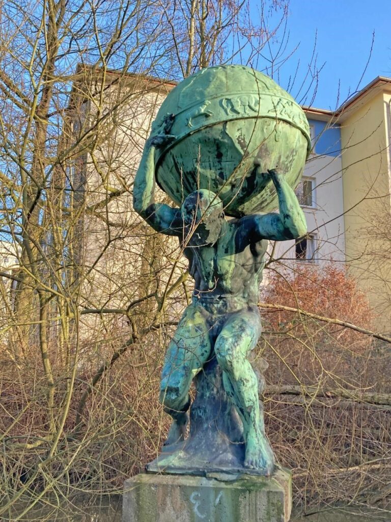 Eine Statue aus Bronze. Ein nackter Mann trägt eine Weltkugel auf seinen Schultern, die er mit beiden Händen festhält.