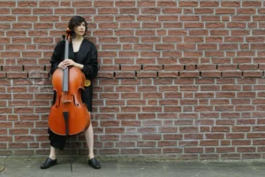 Eine Frau lehnt breitbeinig an einer Backsteinmauer, ihr Cello vor sich.