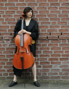 Atena Eshtiaghi steht vor einer Mauer und hält das Cello vor sich.