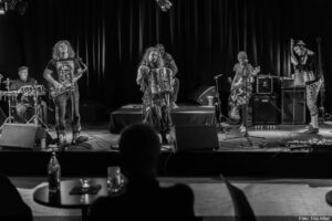 Schwarz-weiß-Bild: 6 Musiker*innen, drei davon mit langen Haaren, zwei mit Kofbedeckungen, stehen mit ihren Instrumenten (Schlagzeug, Saxophon, Akkordeon, Bassgitarre, E-Gitarre) auf der Bühne