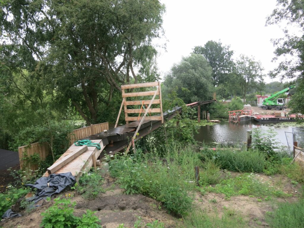 Die Baustelle mit Büschen und in der Mitte ein Steg über die Dove Elbe. Davor eine Absperrung aus Holz.