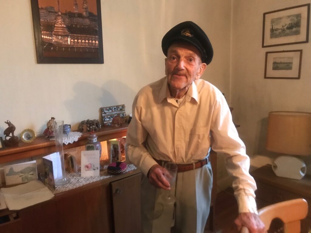 In einem Zimmer. Hinten auf dem Regal Postkarten und Souveniers, in der Mitte Peter Backhausen mit Kapitänsmütze im weißen Hemd und beiger Hose, mit einem Glas in der Hand.