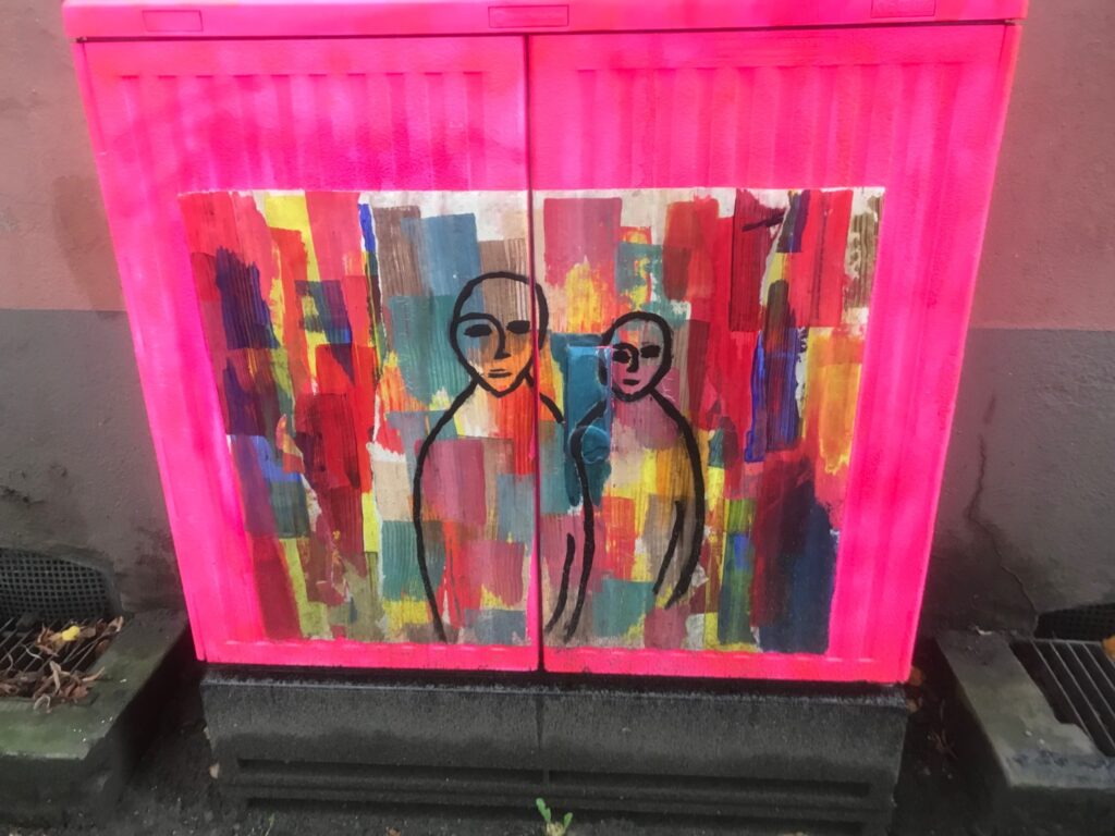 Pinker Stromkasten mit zwei schwarz gezeichneten menschlichen Figuren auf vielfarbigem Hintergrund.