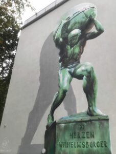 Der Atlasmann als Wandgemälde an einer weißen Hauswand auf einem Sockel. Auf dem steht: "Im Herzen Wilhelmsburger". Die Figur ist grün wie Grünspan von Kupfer.
