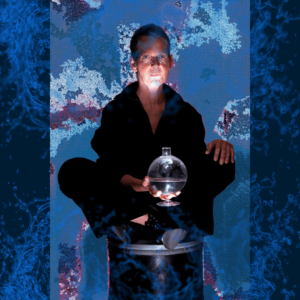 Eine ältere Frau sitzt im Schneidersitz in dunkler Kleidung auf einem Hocker im Schneidersitz, in ihrem Schoß eine Glaskugel, von der sie angeleuchtet wird. Blauer Hintergrund.