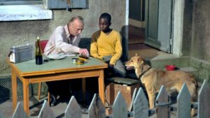 Ein grauhaariger weißer Mann und ein schwarzer Teenager sitzen an einem Tisch im Freien, darauf ein Radio, eine Weinflasche und eine Tabakdose. Ein Hund schaut zu. Ein Zaun im Vordergrund.