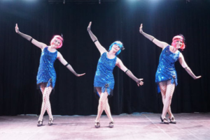 3 Frauen* stehen in synchroner Haltung in blauen, kurzen Glitzerkleidern auf der Bühne.
