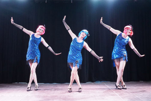 3 Frauen stehen in synchroner Haltung in blauen, kurzen Glitzerkleidern auf der Bühne.