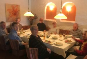 6 Herren und zwei Damen sitzen um einen weiß gedeckten Tisch. Vor ihnen stehen Teller mit Essen und Gläser mit Getränken. 