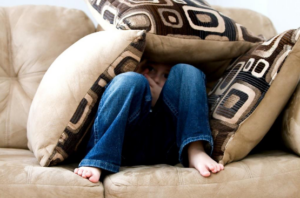 Unter einem Stapel Sofakissen gucken die Beine eines Kindes hervor, das auf einem Sofa sitzt.
