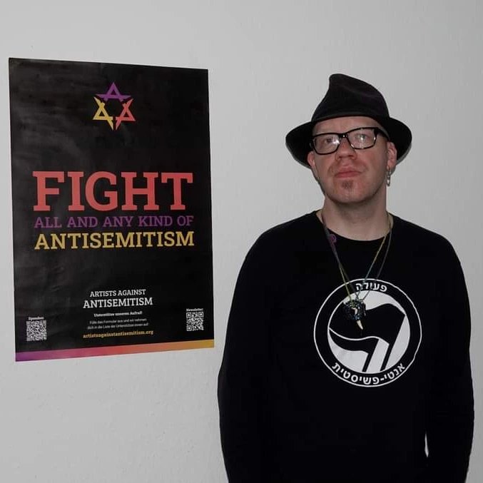Ein Mann um die 40 steht mit schwarzem hut und schwarzem Antifa-Shrt vor einer Wand. Daran hängt ein Plakat, auf dem Steht "Fight all and any kinds of Antisemitism"
