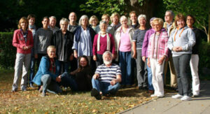 Eine Gruppe älterer Damen und Herren steht und sitzt lächelnd vor Bäumen.